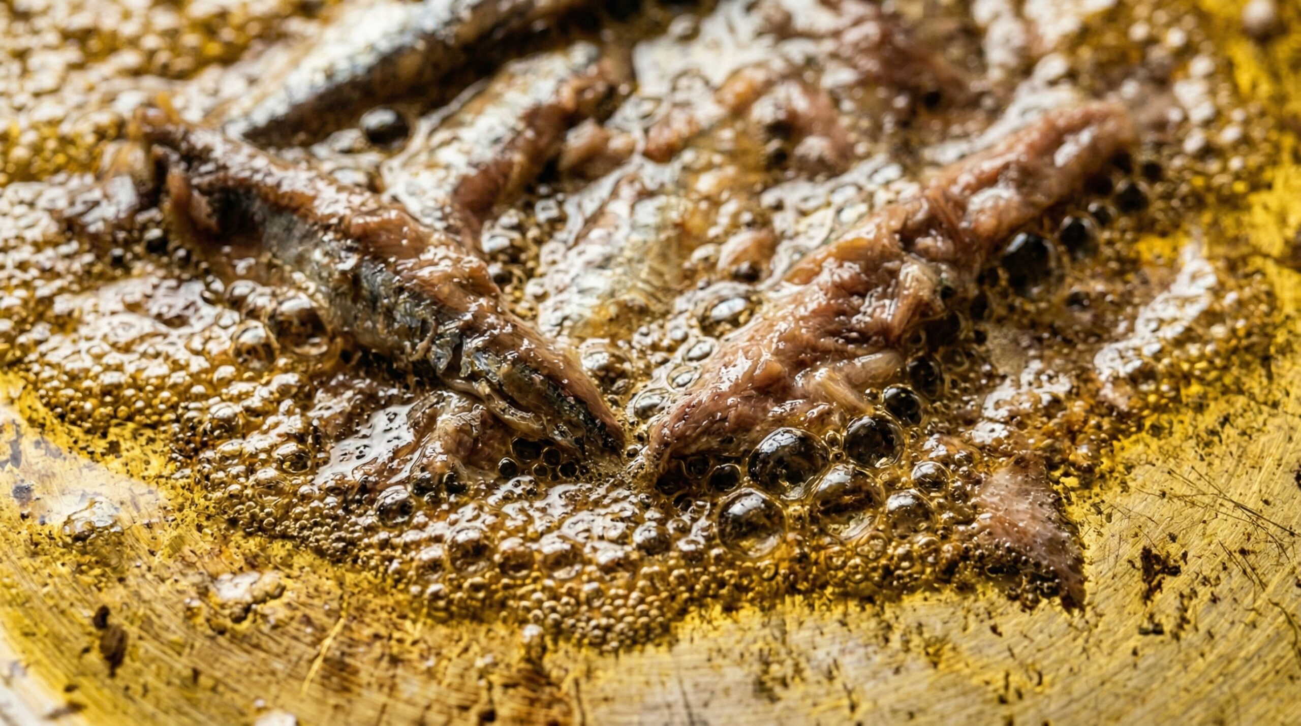 Macro detail of anchovy fillets actively dissolving into a pool of hot, shimmering golden olive oil in a stainless steel skillet