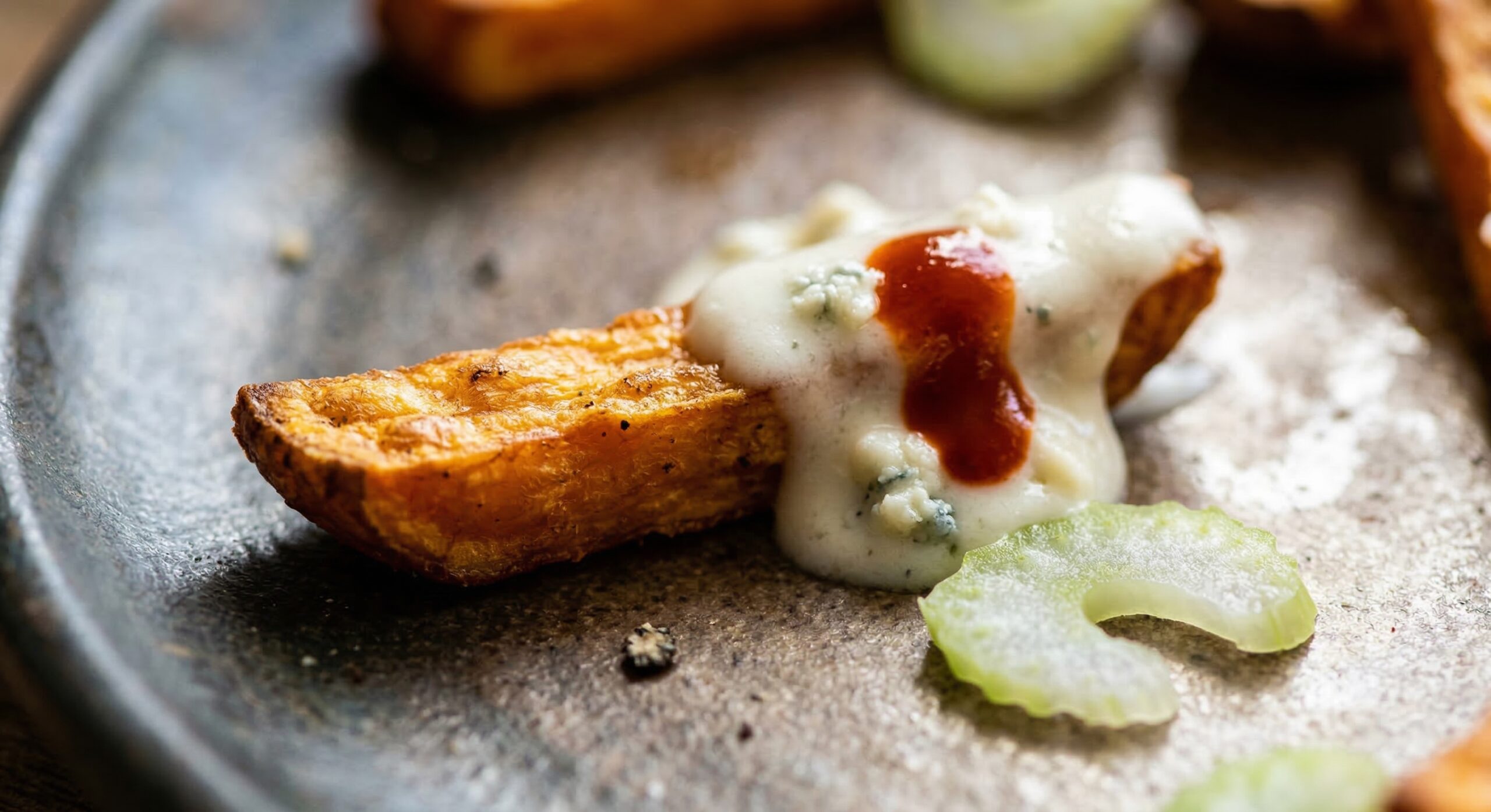 Macro close-up of a golden sweet potato fry enrobed in creamy blue cheese gravy and a drop of bright red hot sauce, with coarse black pepper and a celery slice.
