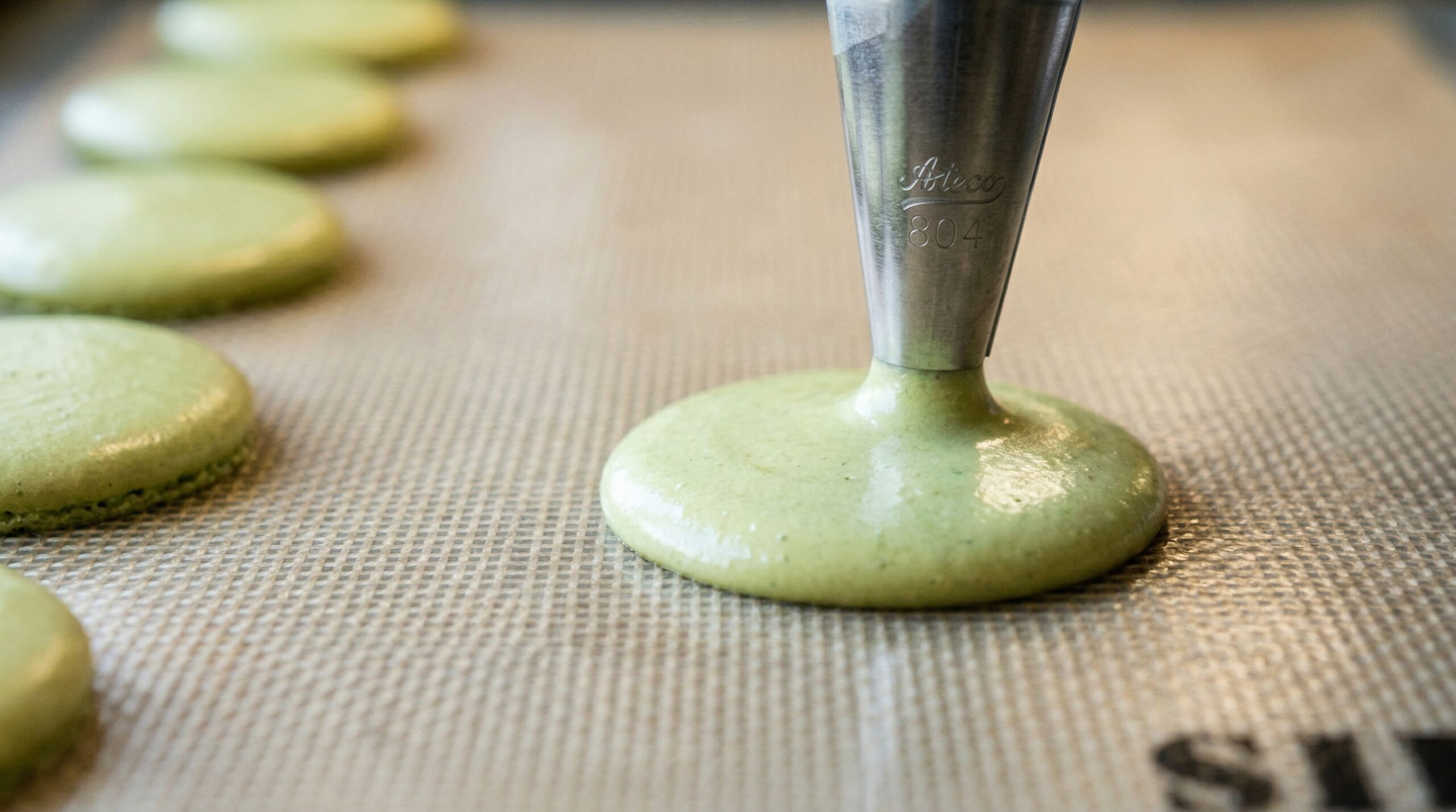 Macro detail of a professional silicone piping bag extruding smooth, round circles of pale green macaron batter onto a Silpat mat