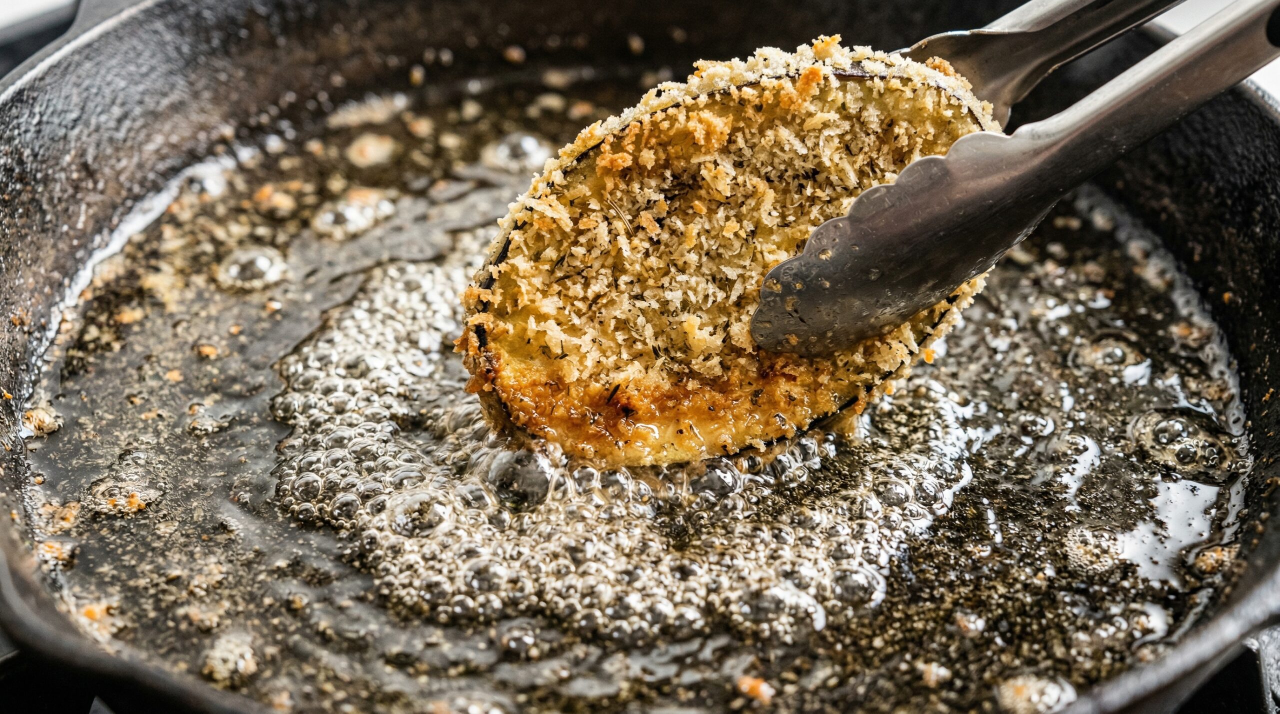 Macro detail of breaded eggplant slices searing in a bubbling cast-iron skillet