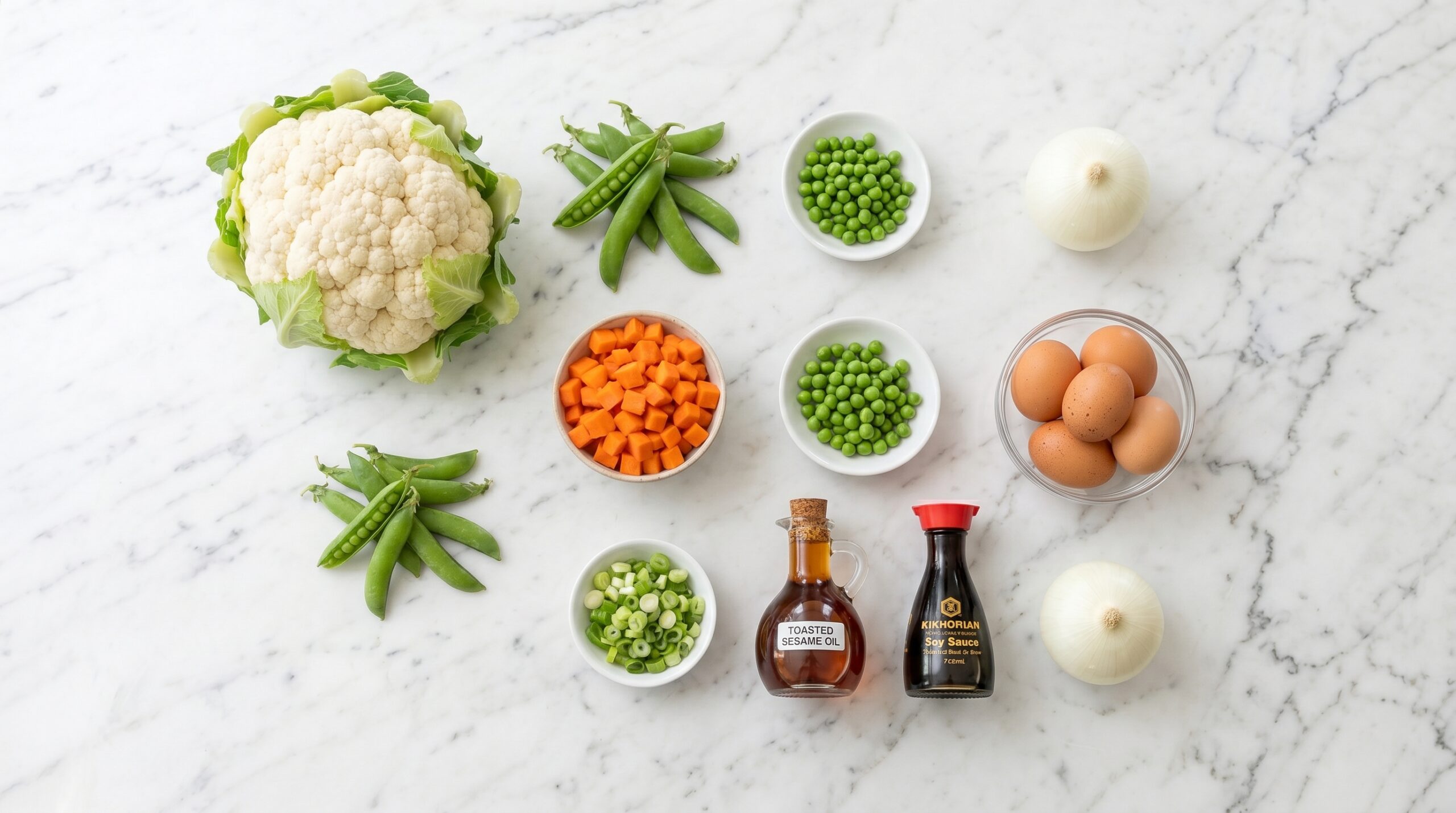 Mise-en-place flat-lay on a marble island showing a whole head of cauliflower, fresh green peas, carrots, white onion, eggs, toasted sesame oil, and soy sauce