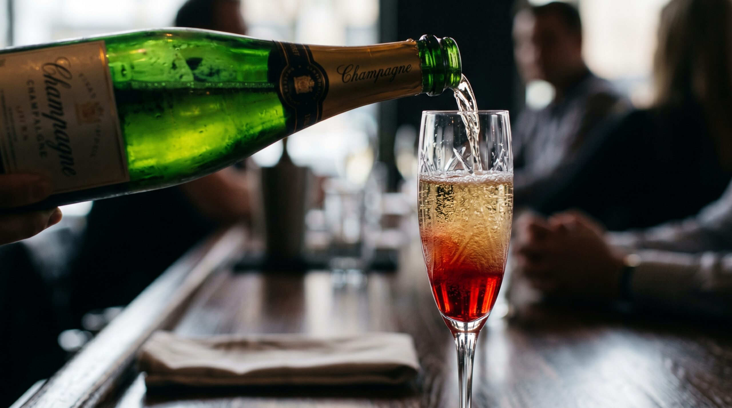 Action shot of sparkling Champagne cascading into a tall crystal flute filled with vibrant red pomegranate and Chambord base