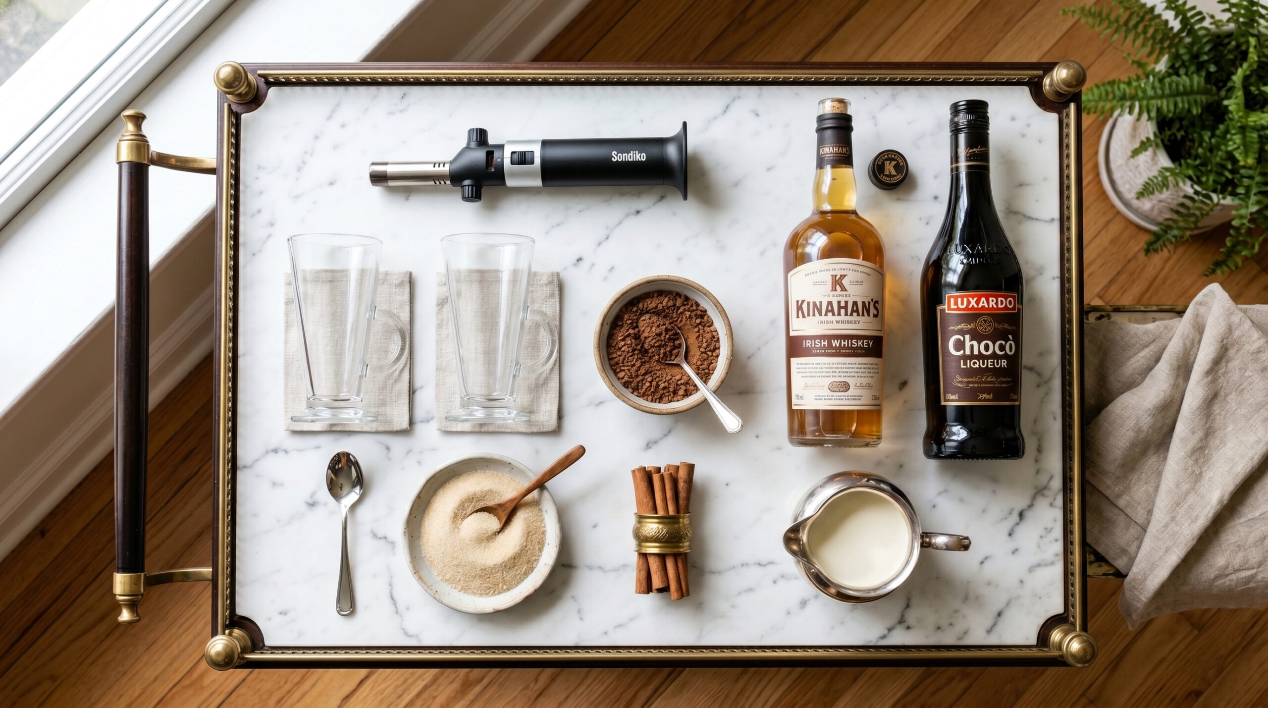 Mise-en-place flat-lay on a marble bar cart showing clear glass mugs, a culinary torch, whole cinnamon sticks, premium whiskey, dark chocolate liqueur, and fresh cream