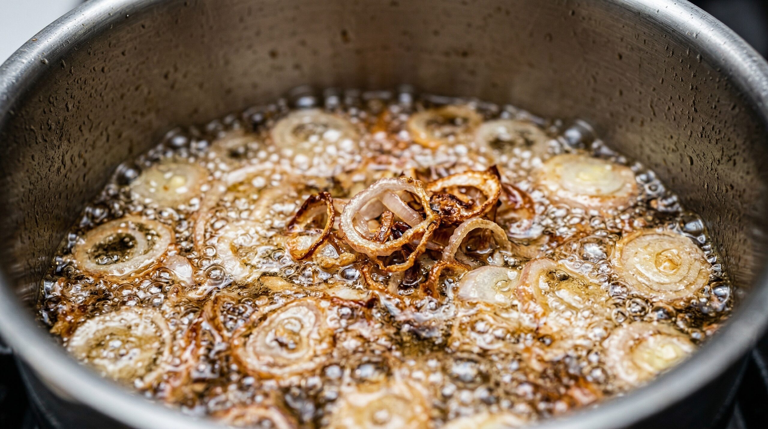 Macro detail of thin rings of raw shallots actively bubbling and rapidly frying in hot canola oil, turning a shattered, crispy golden brown