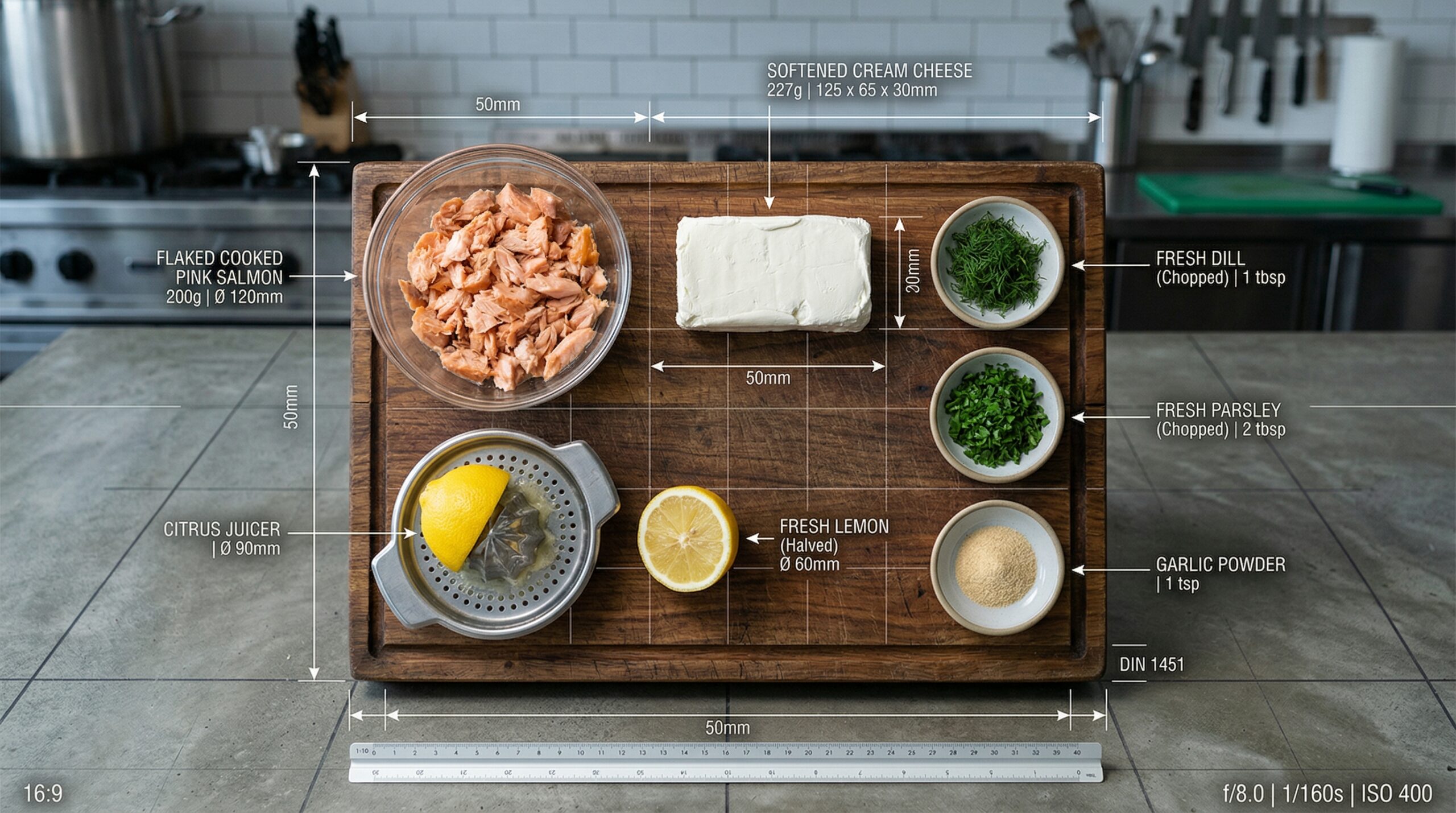 Flaked, cooked pink salmon resting next to a block of softened cream cheese, a silver citrus juicer, and fresh dill on a wooden prep board