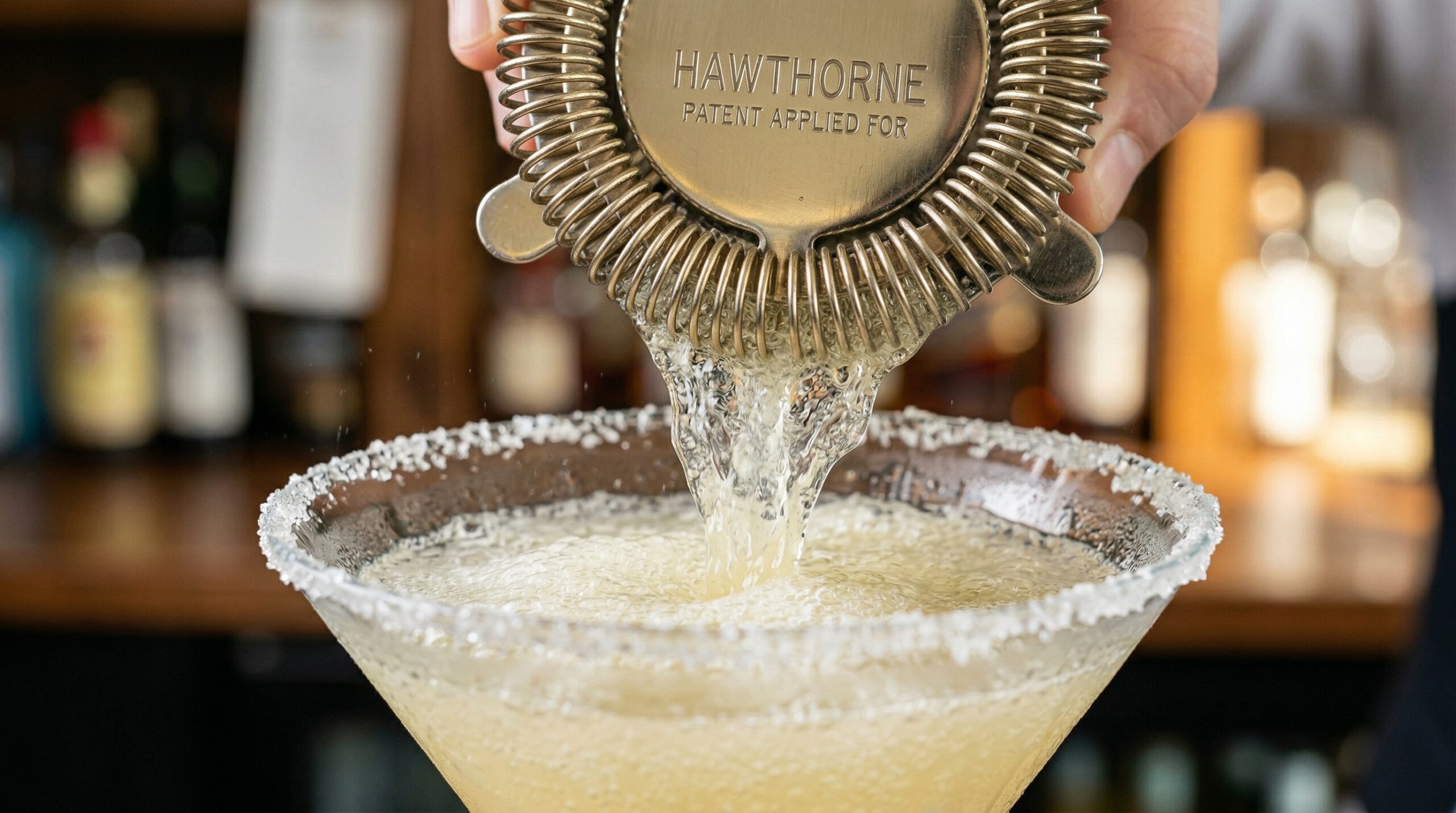 Macro detail of a frosty, ice-cold, pale-yellow cocktail actively pouring through a stainless steel Hawthorne strainer directly into a sugar-rimmed crystal martini glass
